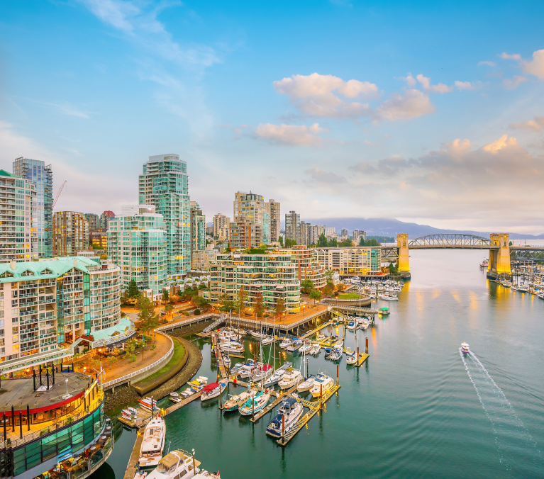 A vibrant view of Vancouver's False Creek at sunset, showcasing modern residential buildings, a marina filled with boats, and the iconic Burrard Street Bridge in the background. The calm waters and warm light create a picturesque and inviting urban waterfront scene.